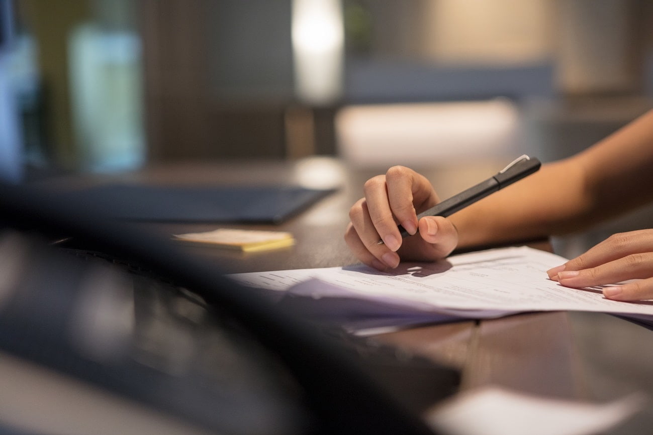 Close-up of a hand holding a pen and reviewing documents on a desk, symbolizing the detailed review and preparation of Pharma SOP and CMC documentation in a professional setting.