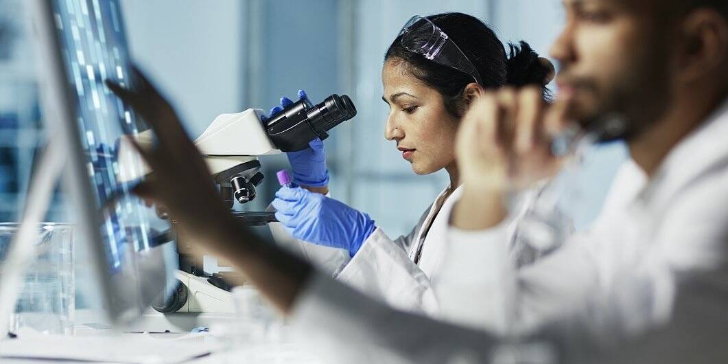 Researchers in a laboratory setting, with a focus on a scientist using a microscope while another analyzes data on a computer screen, symbolizing collaborative efforts in clinical trial documentation and research.