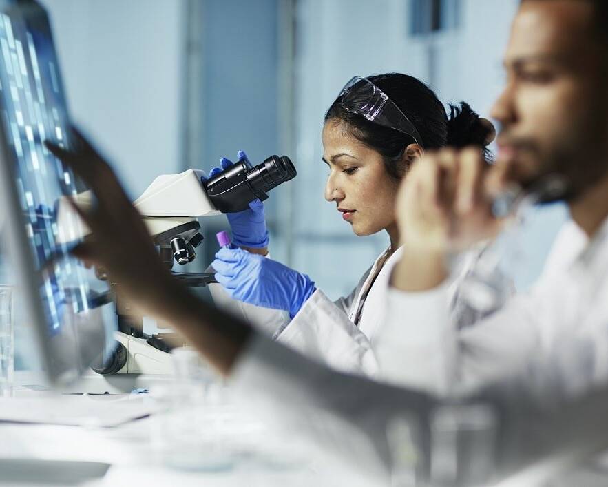 Researchers in a laboratory setting, with a focus on a scientist using a microscope while another analyzes data on a computer screen, symbolizing collaborative efforts in clinical trial documentation and research.