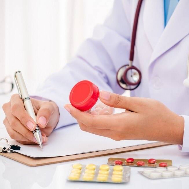 Doctor in a white coat with a stethoscope holding a medication bottle while writing on a clipboard, with various pills and blister packs on the table, representing healthcare practices and global labelling for medication safety and clarity.