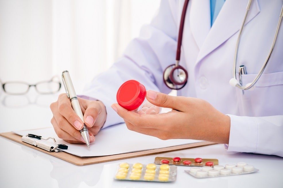 Doctor in a white coat with a stethoscope holding a medication bottle while writing on a clipboard, with various pills and blister packs on the table, representing healthcare practices and global labelling for medication safety and clarity.